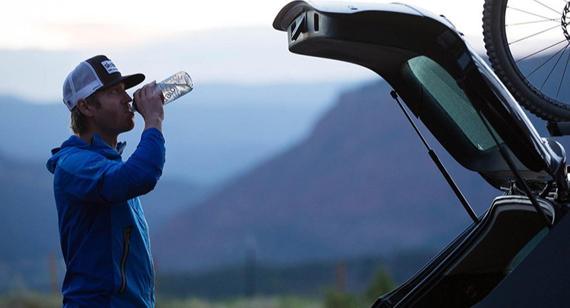 A man drinking water outside next to an open hatchback