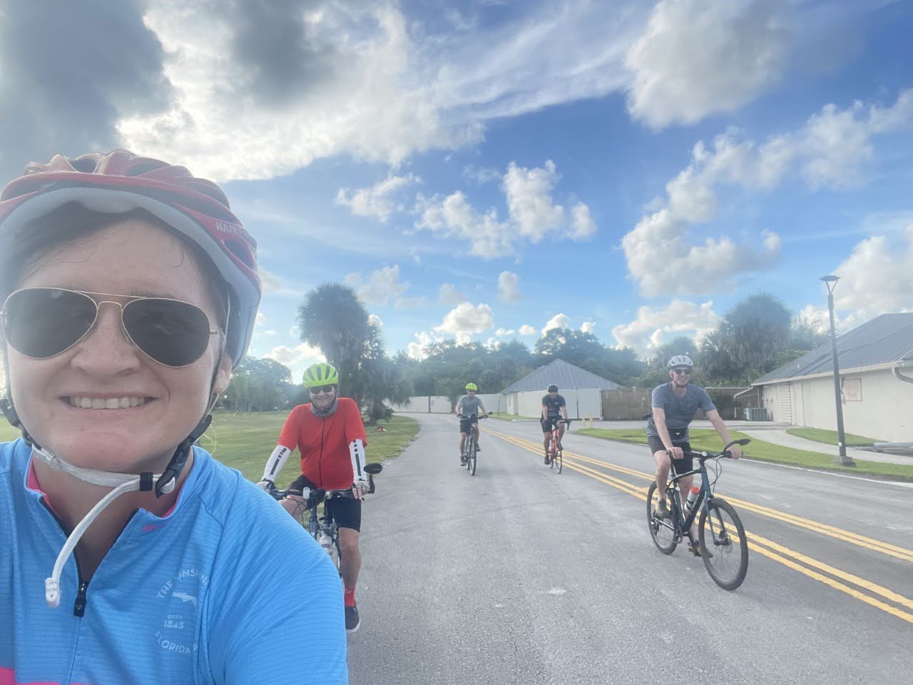 A woman takes a selfie while riding a road bike. 3 other bike riders are in the background on a sunny day.