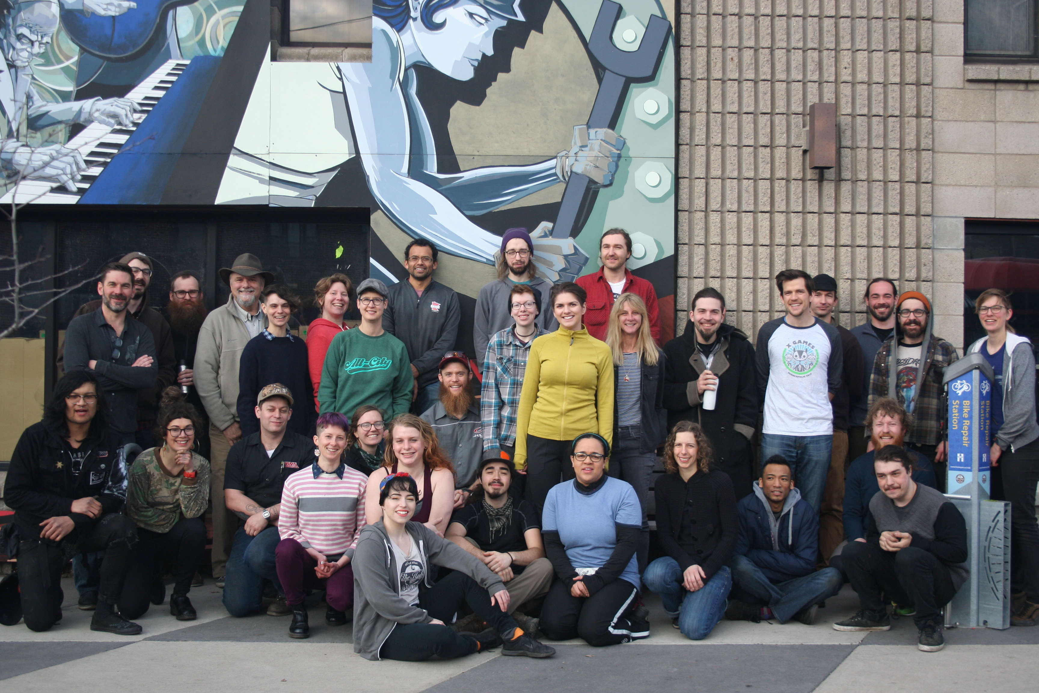 2019 Hub staff photo: group of people against Longfellow shop location with mural facade and concrete brick wall behind