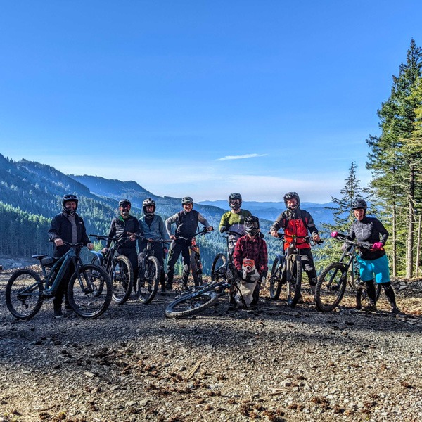 Several mountain bikers and dog posing for a photo with mountains in the background