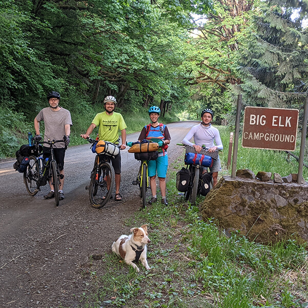 Four cyclists and a dog posing for a photo on their bikes next to a sign that reads "Big Elk Campground"