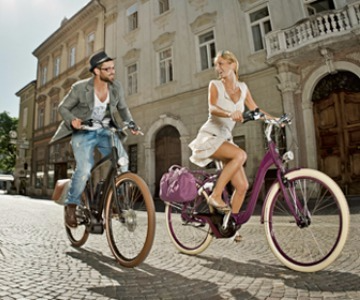 A man and woman riding e-bikes in an old city street.