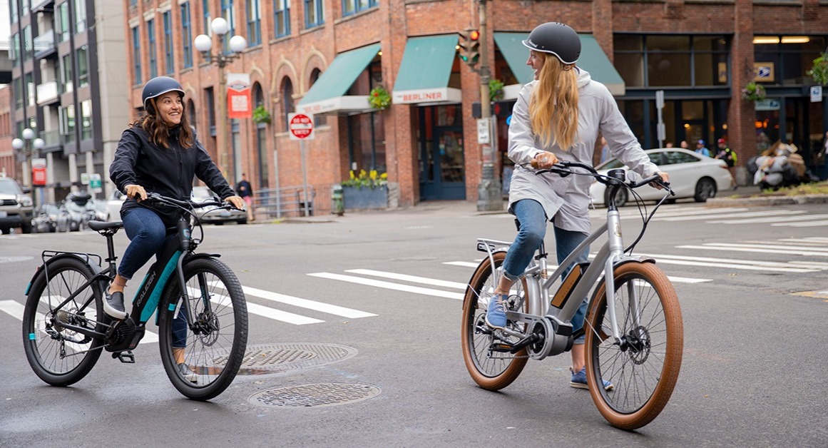 Two women ride Raleigh e-bikes on a city street