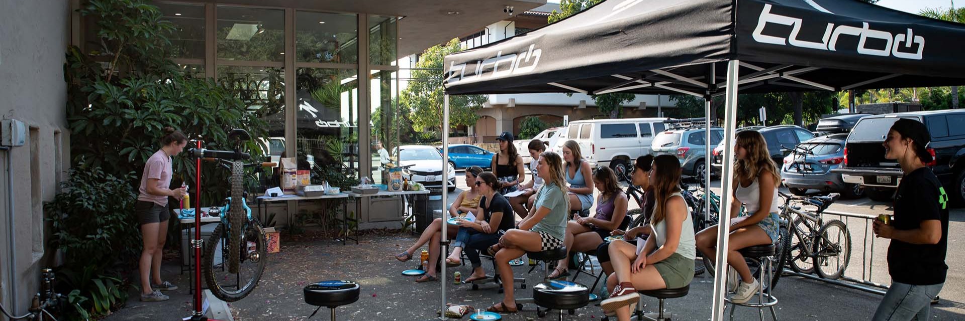 A group of women from the mountainadoras riding group sit under a pop up tent and watch a mechanic at a table filled with tools