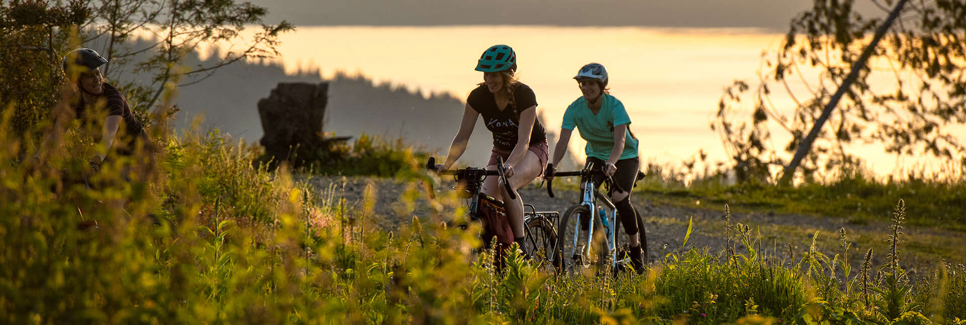 2 girls & 1 guy riding drop bar gravel bikes near lake and mountains at sunset