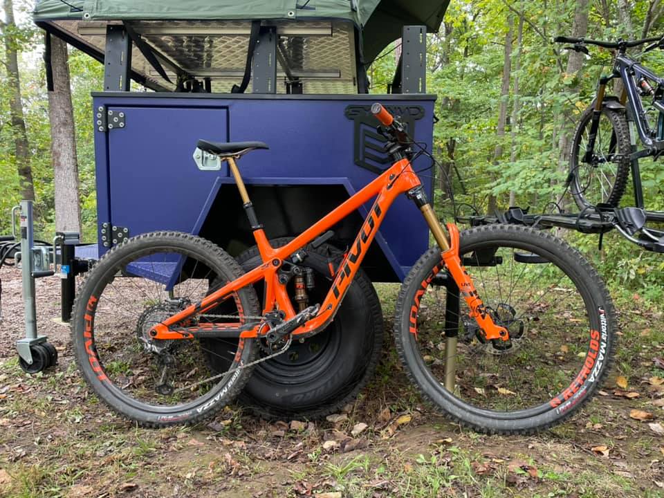 An orange Pivot Firebird leans up against a purple overland trailer.