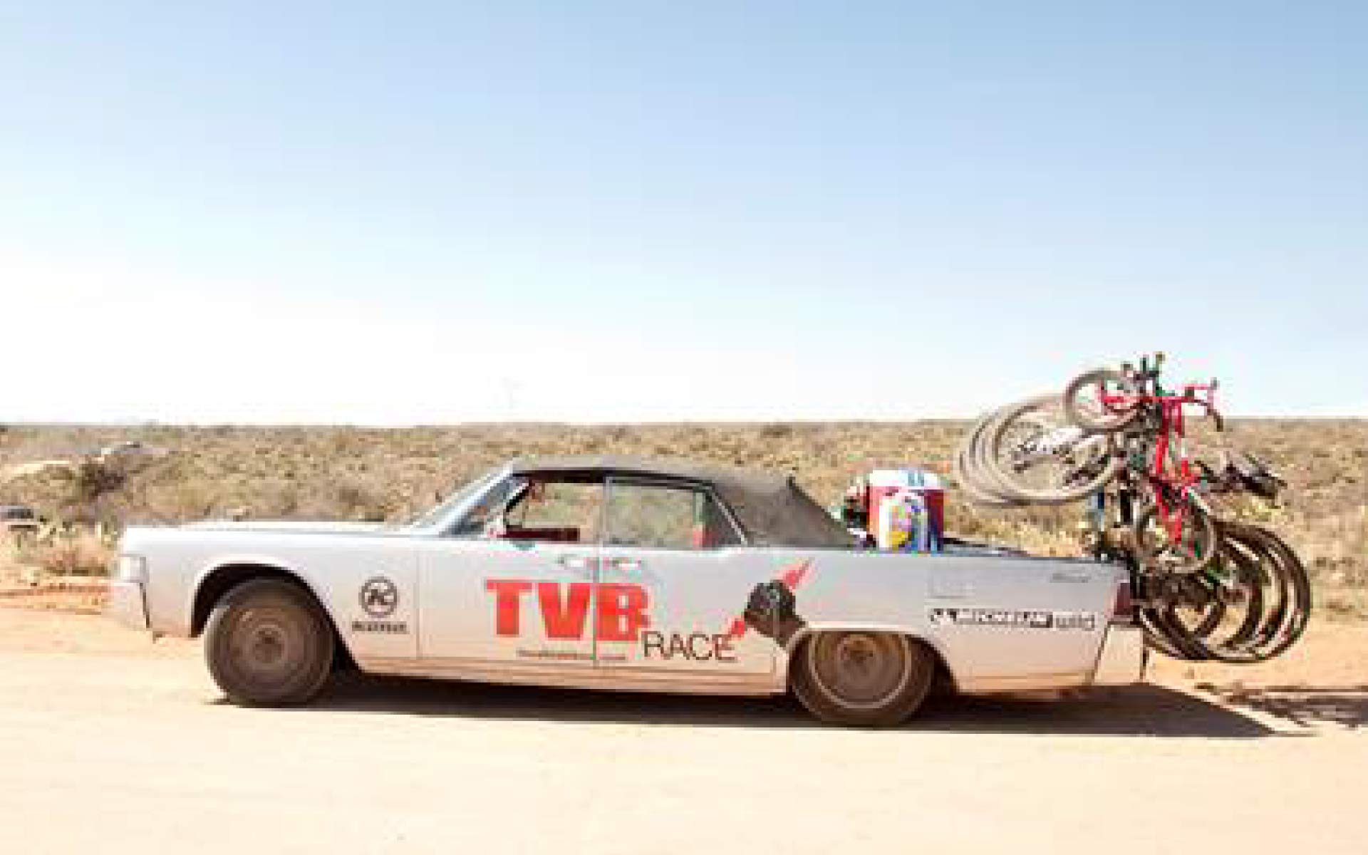 Vintage Car with "TVB Race" on the side drives down a dirt road with bikes mounted on the back