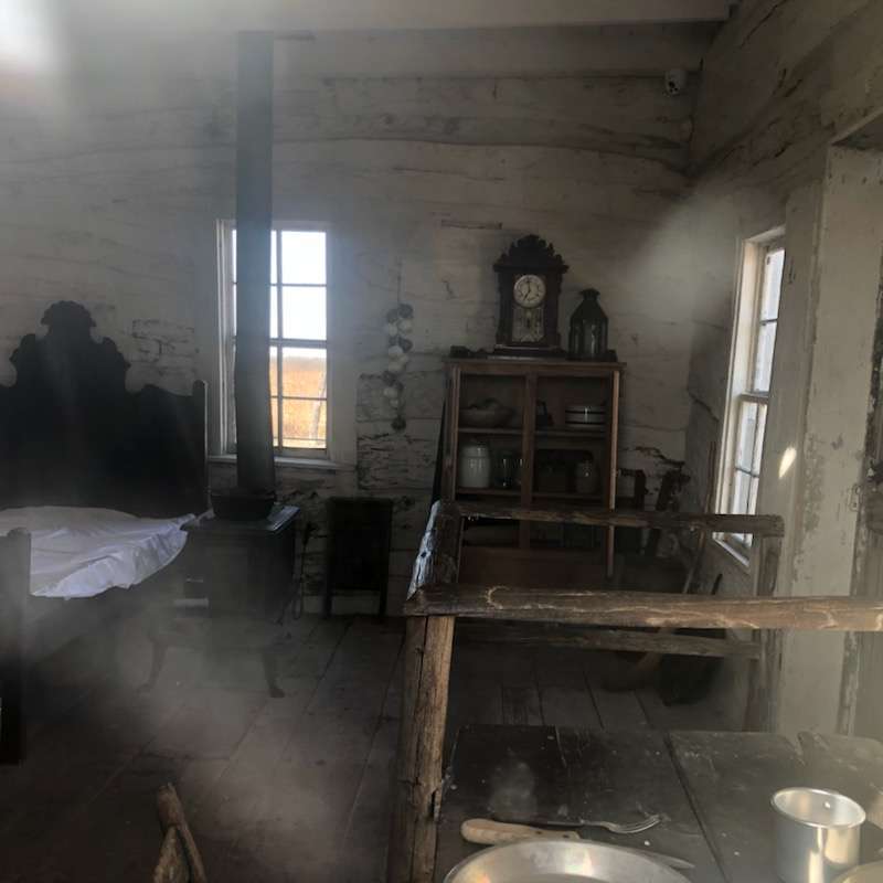 Inside look of homestead: Bed with thin mattress, small wood burning stove with a small shelf with some dishes and a clock with a view of the table with tin dishes