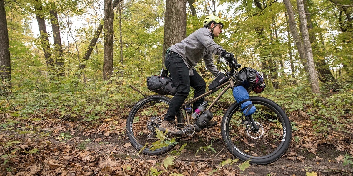 A cyclist rides their bike loaded with camping gear on a dirt trail in the woods.