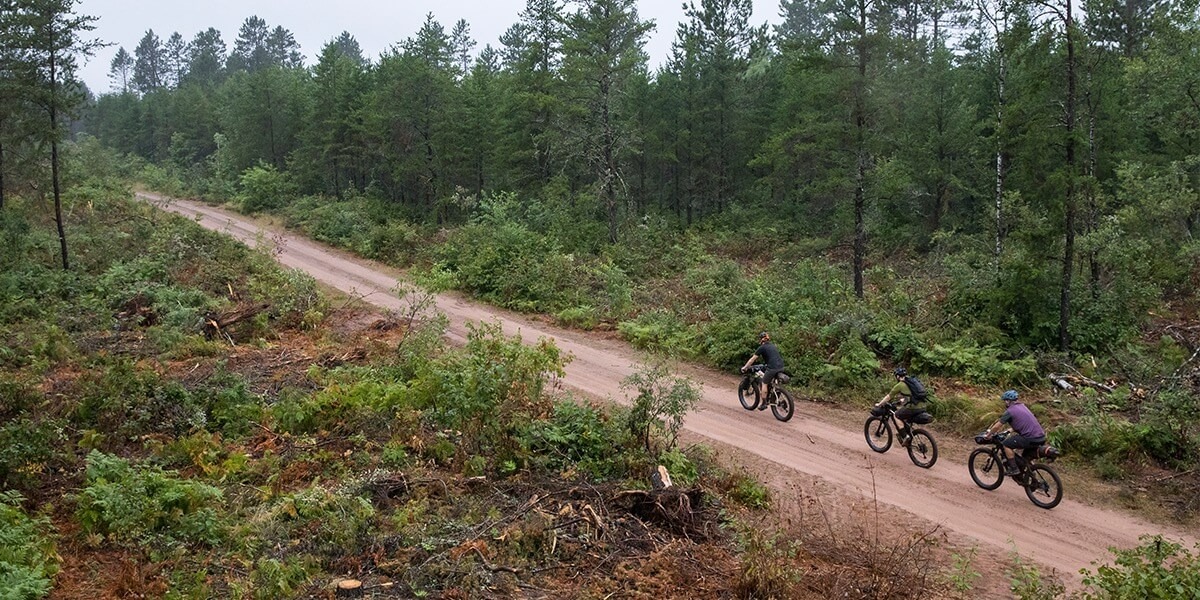 Aerial view of 3 cyclists riding on a dirt road with woods on either side.