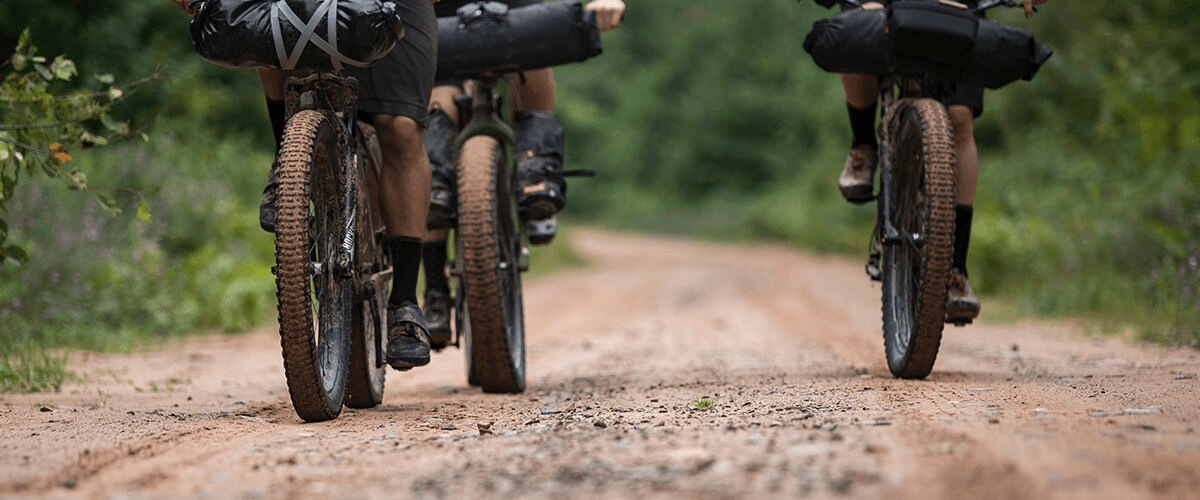 Three cyclist on bikes shown from the tires down on a gravel road riding through the woods.