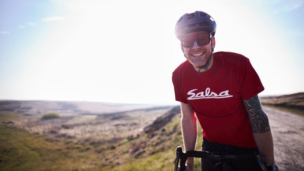 Steve Bate smiles for the camera while standing with his gravel bike in front of grassy peaks and valleys