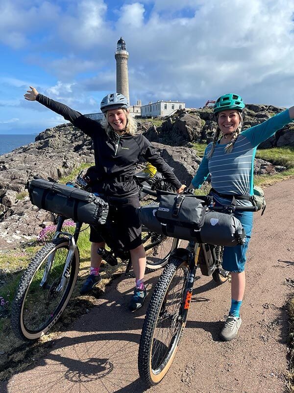Kate and Shophie stand with their bikes loaded with gear in front of a lighthouse on the coast