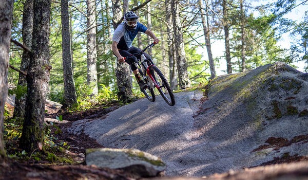 A mountain biker banks a turn and catches air over a rock on a mountain trail.