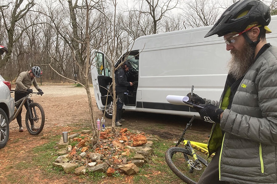 Three cyclists stand near a van and inspect bikes and take notes.