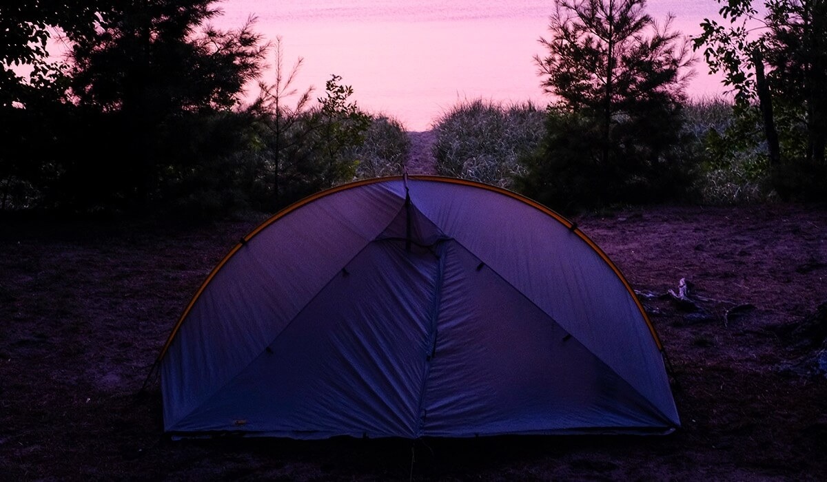 A camping tent is set up on the ground beneath a pink and purple sky with trees in the background.