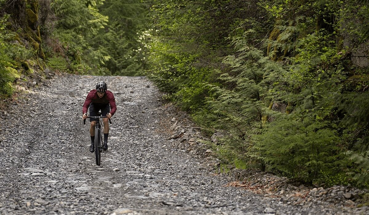 Chris rides his gravel bike on a descent down a rocky path lined with tress.