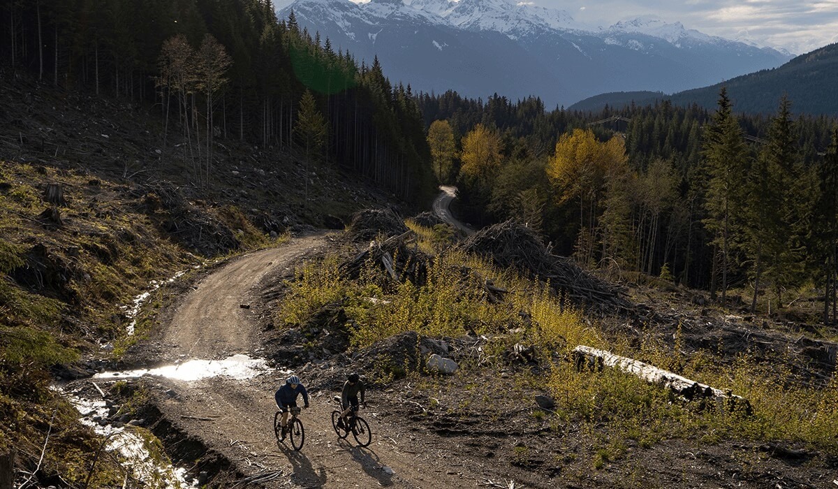 Two cyclists ride side by side on a dirt path through the mountains while the sun shines down.