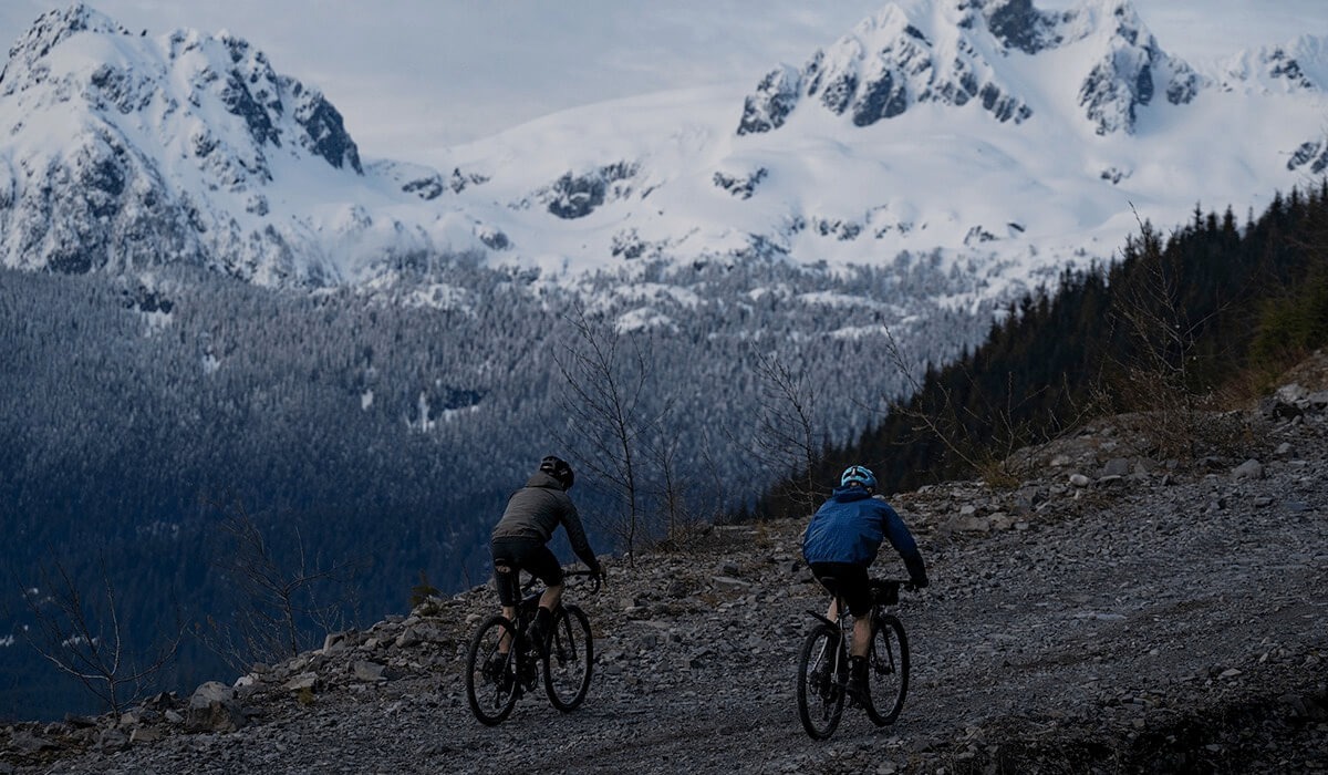 Two cyclists ride together on a rocky trail up a hill with snow covered mountains in the background.