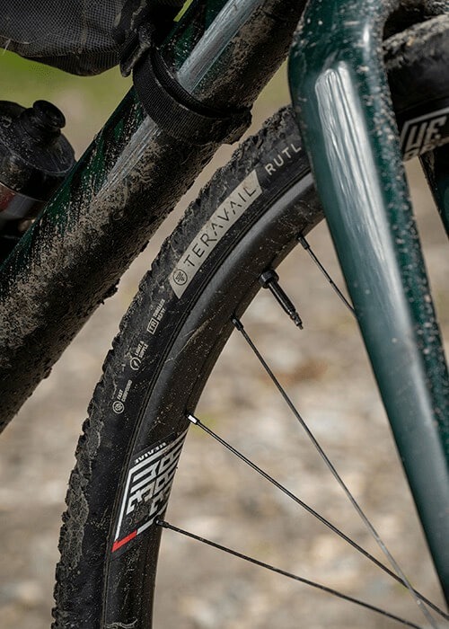 2 images side by side. Left is an aerial view of 2 cyclists on a dirt road. Right is a closeup of a muddy teravail tire and fork