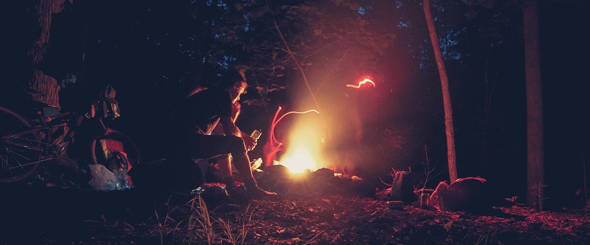 A group of cyclists sit around a campfire in the woods at night.