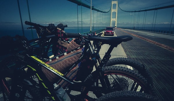 Several mountain bikes are lined up in the foreground on a suspension bridge over water.