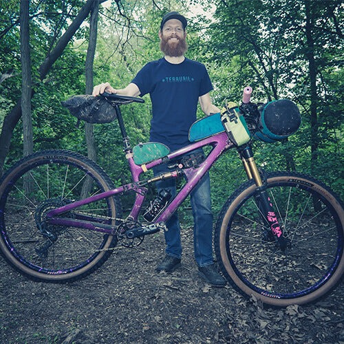 A cyclist poses in the woods with a loaded purple full suspension mountain bike