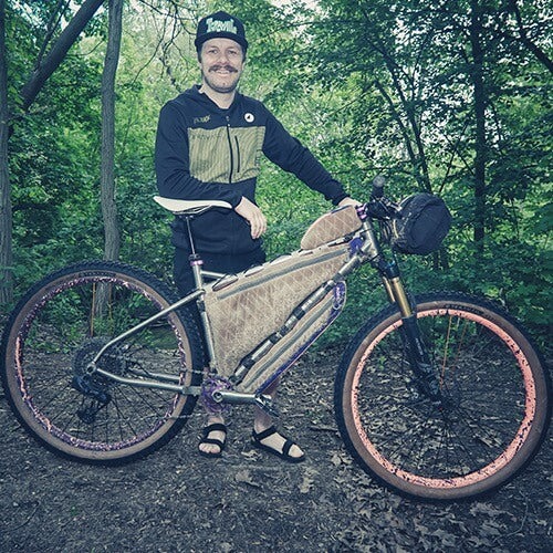 A cyclist poses in the woods with a loaded tan hardtail mountain bike
