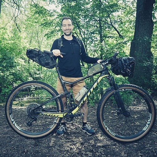 A cyclist poses in the woods with a loaded black and yellow full suspension mountain bike