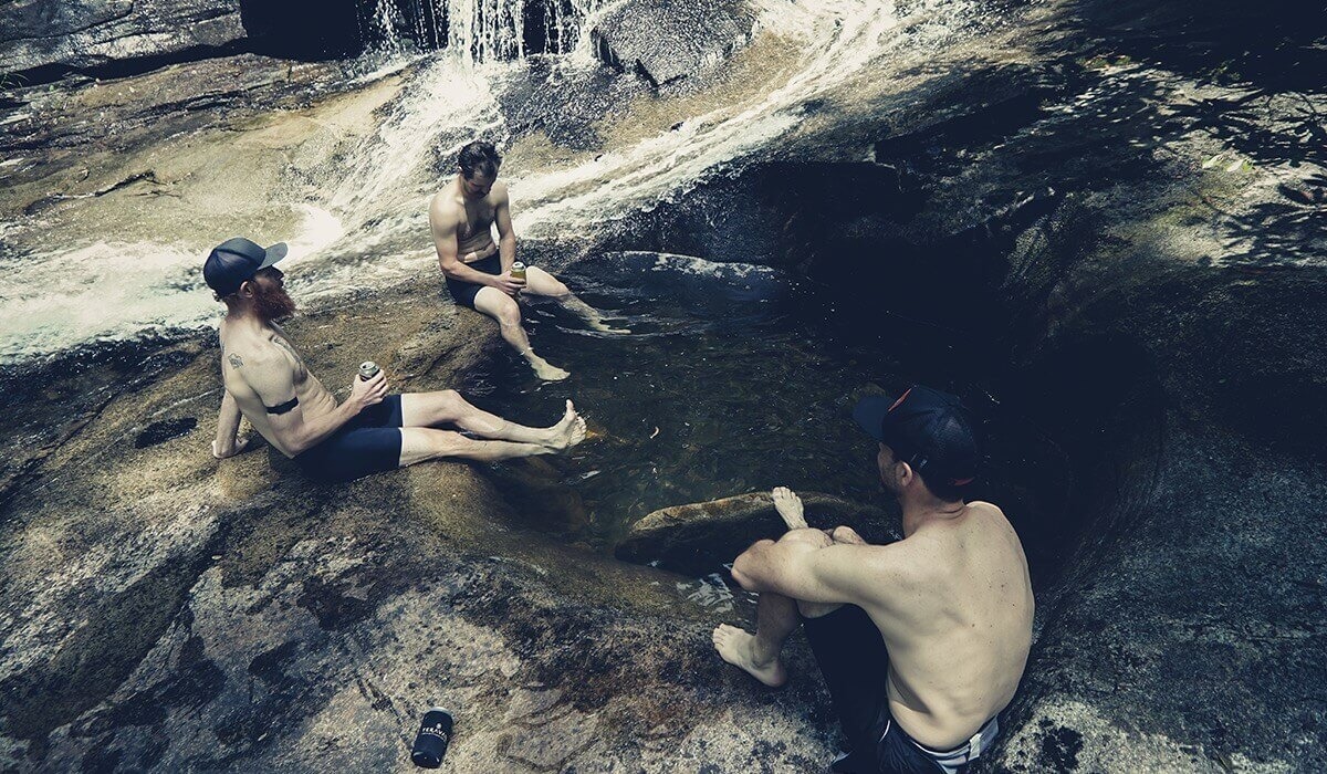 Three riders relax and cool down with drinks at the rocky bottom of a water fall