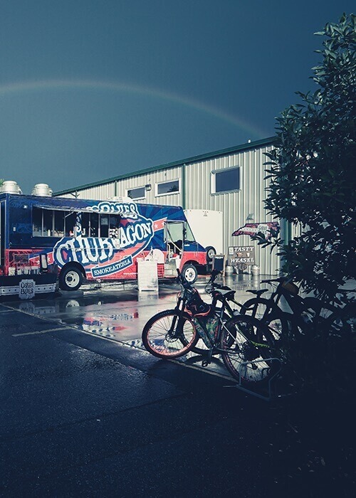Bikes are parked outside of a brewery. There is a rainbow in the sky.