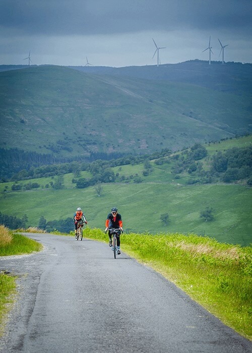 Two cyclists ride on a narrow paved road through a hilly landscape.