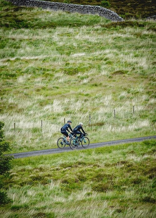 Two cyclists ride side by side on a paved road that runs through a grassy hillside