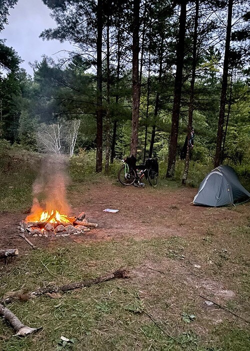 Adam's campsite has a tent, fire, and his bike leaning against a tree