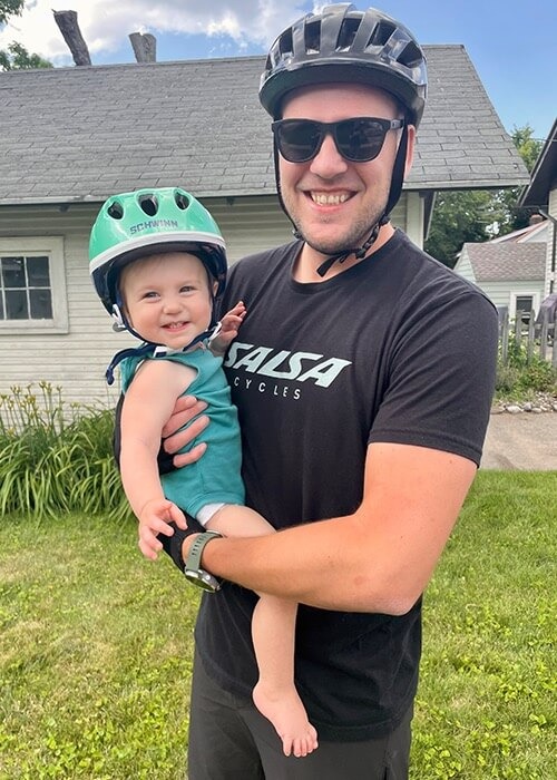 Adam hold his daughter and smiles for the camera. They both have their bike helmets on.