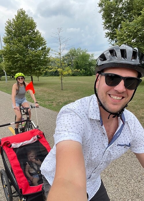 Adam and his wife ride on a paved bike path with their daughter in a bike trailer