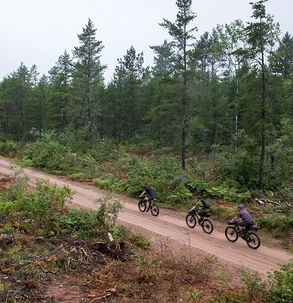 Three cyclists shown from afar ride in a line on a dirt road on mountain bikes packed with gear.