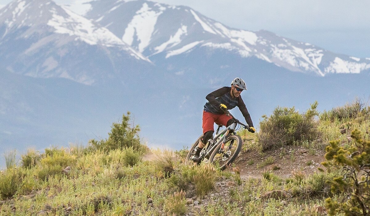 A mountain bike rides through a flat narrow dirt trail with a snow capped mountain in the background.