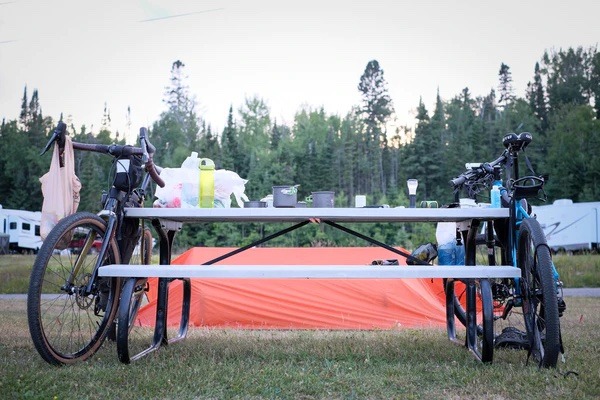 2 bikes are leaning against a picnic table with camping gear
