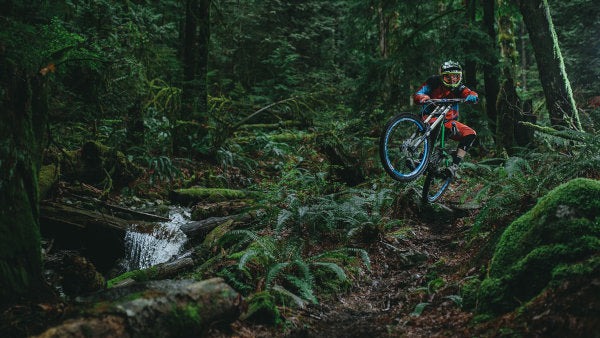 Kyle Quesnel doing a wheelie on his mountain bike through a lush forest trail