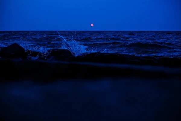 Waves from Lake Superior crash into the shore at night while the moon shines in the sky.