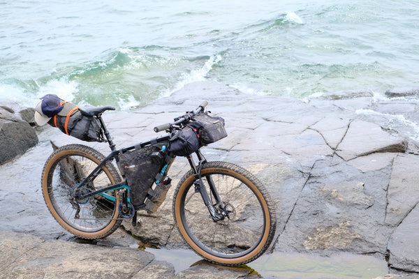 A bike loaded with camping gear sits on a rock at the lake's edge while waves come in.