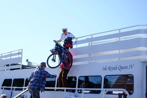 The captain of the ferry lifts a loaded gravel bike down over the side of the boat to the riders