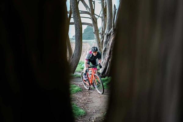 A cyclist is seen through the trees riding a gravel bike on a dirt trail