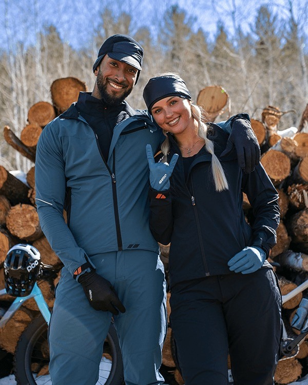 Two riders stand together in front of a log pile wearing their 45NRTH Naughtvind jackets and winter cycling gear.