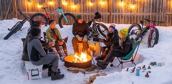 A group of cyclists sit around a fire pit. A mound of snow and several fat tire bikes are in the background.